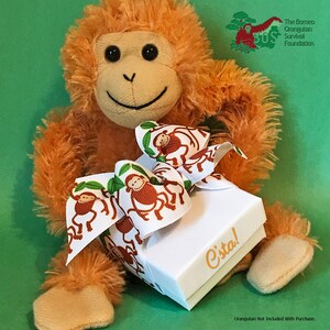 May include: A plush orange orangutan holding a white gift box with a ribbon tied in a bow. The ribbon is decorated with a pattern of monkeys swinging from vines. The box has the text "C'sta!" printed on the top.
