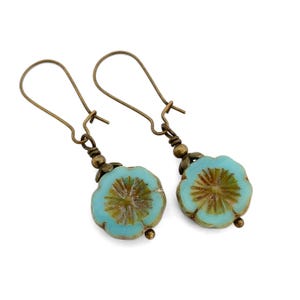 May include: A pair of handcrafted earrings featuring light blue flower-shaped beads with brown centers. The earrings have antique bronze-colored ear wires and decorative accents. The floral beads are detailed with etched patterns, creating a vintage aesthetic.
