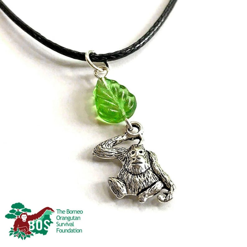May include: A silver orangutan charm necklace with a green glass leaf pendant. The necklace is on a black cord. The charm is a depiction of an orangutan sitting with its arm raised. The necklace is a fundraiser for The Borneo Orangutan Survival Foundation.
