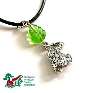 May include: A silver orangutan charm necklace with a green glass leaf pendant. The necklace is on a black cord. The charm is a silhouette of an orangutan hanging from a branch. The necklace is a fundraiser for the Borneo Orangutan Survival Foundation.