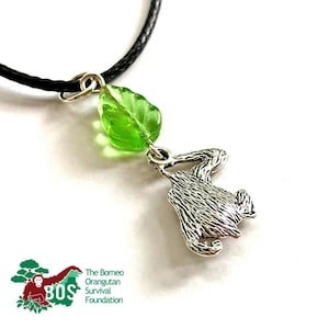 May include: A silver orangutan charm necklace with a green glass leaf pendant. The necklace is on a black cord. The charm is of an orangutan holding a leaf in its hand. The necklace is a fundraiser for the Borneo Orangutan Survival Foundation.