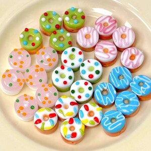 May include: Assortment of colourful glass donut beads with various designs and sprinkles. The beads are pink, blue, green, white, and yellow.