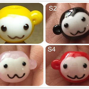 May include: Four glass monkey beads in different colors: yellow, black, pink, and red. Each bead has a white face with black eyes and a smiling mouth.