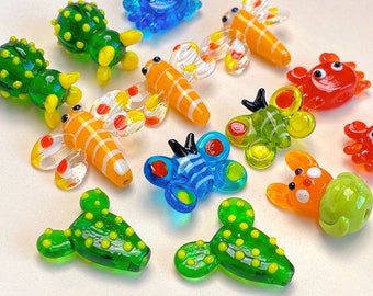 Puede incluir: Una colección de abalorios de animales e insectos de vidrio de colores, que incluyen un cangrejo azul, un cactus verde, una libélula amarilla y naranja, una mariposa azul y una mariquita roja.