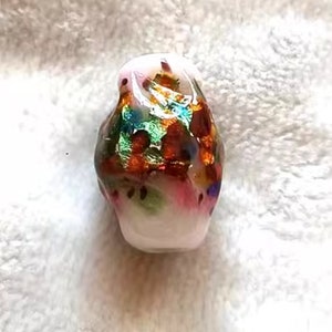 Puede incluir: Una cuenta de vidrio blanca con un diseño colorido e iridiscente. La cuenta tiene una superficie texturizada y presenta tonos de naranja, verde, azul y rosa.