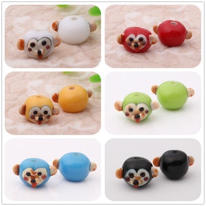 May include: Set of 10 colorful glass monkey beads. Each bead features a monkey face with brown eyes, a red nose, and a pink tongue. The beads are available in white, red, yellow, green, blue, and black.