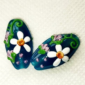 May include: Two teal blue glass beads with white flowers and green leaves. The beads are oval shaped and have a glossy finish.