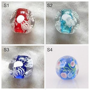 May include: Four glass beads with different designs. The first bead is red with a white jellyfish inside. The second bead is blue with a white jellyfish inside. The third bead is blue with a white jellyfish inside. The fourth bead is blue with pink and green designs.