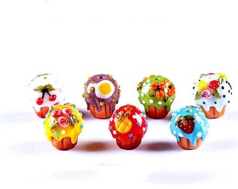 May include: A collection of eight colorful glass cupcake beads with various toppings, including cherries, flowers, pineapple, and strawberries. The beads are all approximately the same size and are made of clear glass with painted details.