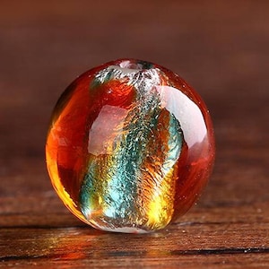 May include: A round glass bead with a swirling pattern of orange, gold, and teal colors. The bead has a shiny, reflective surface.