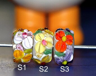Peut inclure: Trois perles de verre avec des motifs floraux. La perle de gauche a un fond blanc avec des fleurs roses, jaunes et vertes. La perle du milieu a un fond blanc avec des fleurs jaunes. La perle de droite a un fond violet avec des fleurs orange.