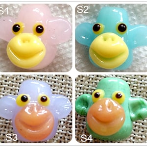 May include: Four glass monkey bead charms in different colors: pink, blue, purple, and green. Each bead has a yellow smile and black eyes.