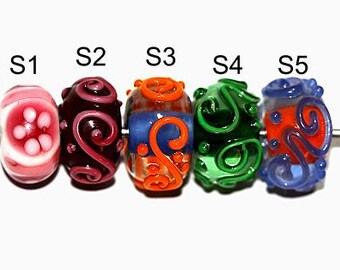 Peut inclure: Cinq perles en verre de différentes couleurs et motifs. Les perles sont disposées en rangée de gauche à droite. La première perle est rose avec des points blancs, la deuxième perle est bordeaux avec un motif en spirale, la troisième perle est bleue et orange avec un motif en spirale, la quatrième perle est verte avec un motif en spirale, et la cinquième perle est bleue avec un motif en spirale orange.