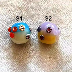 May include: Two glass beads with colorful designs. The bead on the left is blue, white, and orange. The bead on the right is purple, yellow, and white.