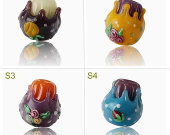 May include: Four glass beads with different colors and designs. The beads are purple, yellow, orange, and blue. Each bead has a different design, including flowers, dots, and swirls.