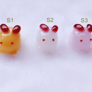 以下が含まれることがあります： 赤い耳と目で、3つの小さなカラフルなウサギの形をした置物。左のウサギは黄色、真ん中のウサギは白、右のウサギはピンク。ウサギはS1、S2、S3とラベル付けされています。