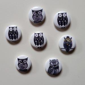 Op de afbeelding: Zeven witte, ronde knopen met zwart-witte uilenontwerpen. De uilenillustraties variëren, sommige met hoeden of brillen. De knopen zijn gerangschikt op een lichtgekleurd oppervlak, met verschillende uilenmotieven.