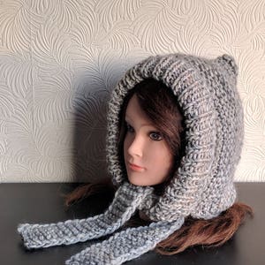 Può includere: Un cappuccio lavorato a maglia grigio e bianco con lacci per sciarpa attaccati. Il cappuccio presenta una parte superiore a punta ed è realizzato con filato spesso e strutturato. I lacci della sciarpa pendono su entrambi i lati. Il cappuccio è esposto su una testa di manichino.