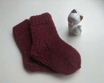 May include: A pair of knitted burgundy socks. The socks are made of a soft, textured yarn and have a ribbed cuff. A small, white ceramic cat figurine with gold accents is in the background.