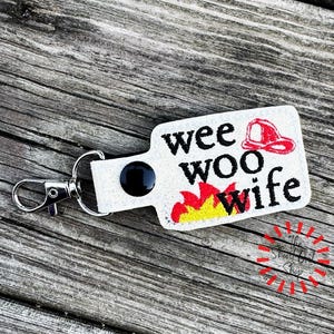 May include: A white rectangular keychain with the words "wee woo wife" in black text, a red firefighter's helmet, and flames in red and yellow. The keychain has a silver clasp and a black circular accent.