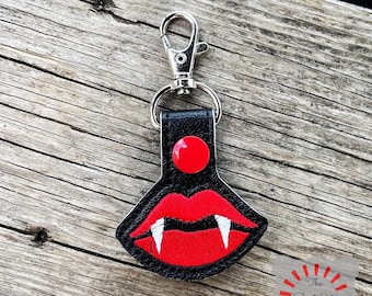May include: A black leather keychain with a red and white vampire mouth design. The keychain has a silver metal clasp.