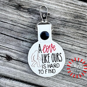 May include: White round keychain with the text "A love like ours is hard to find" in black and red. A Bigfoot silhouette is also featured. The keychain has a silver clasp and a black button.