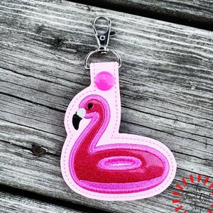 Peut inclure: Un porte-clés en forme de flamant rose avec un motif brodé de couleur rose foncé vif et un bec noir et blanc. Le porte-clés est fixé à un mousqueton en métal argenté et comprend un petit bouton-pression de couleur rose.