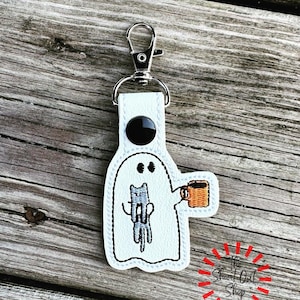 May include: A white ghost-shaped keychain with a black button and silver clasp. The ghost has a cat design and holds a mug. The keychain is on a wooden surface. The text "The Sweet Owl Shop" is in a red burst.