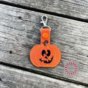 May include: Orange pumpkin-shaped keychain with a black stitched-on face. The keychain has a silver metal clasp and a small orange button.
