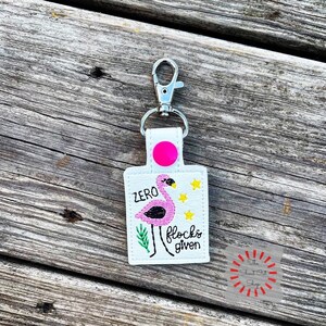 May include: White keychain with a pink flamingo design and the text "ZERO flocks given." The keychain has a silver clasp and a pink circular accent. The flamingo is pink and black with yellow star accents.