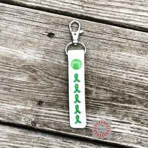 May include: White keychain with a green ribbon design and a silver clip. The ribbon is embroidered on the keychain.