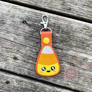 May include: A Halloween-themed keychain featuring a candy corn design with a smiling face. The keychain has a yellow, orange, and white candy corn shape with a silver clasp and orange leather strap. The background is a weathered wooden surface.
