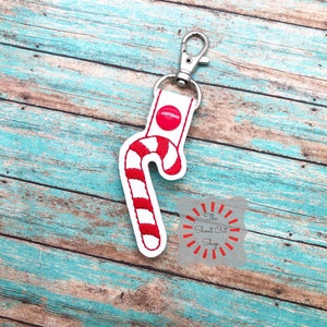 May include: White felt candy cane keychain with red stripes and a red button. The keychain has a silver metal clasp. The text "The Shout Out Shop" is visible in the background.
