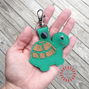 May include: Green leather turtle keychain with a brown shell and black button eyes. The keychain has a silver metal clasp. The text "The Shout Out Shop" is visible in the lower right corner of the image.