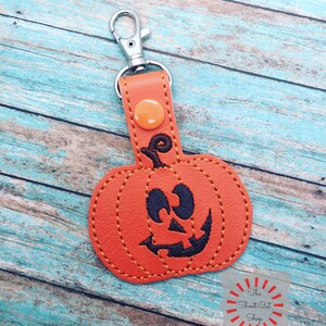 May include: Orange leather pumpkin keychain with a black stitched face and a silver metal key ring. The keychain is on a light blue wooden background. The text "The Shout Out Shop" is visible in the lower right corner of the image.