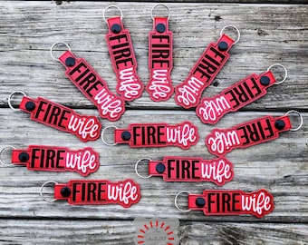 May include: Twelve red keychains with black lettering that reads "FIRE wife" in a white cursive font. The keychains are scattered on a wooden surface.