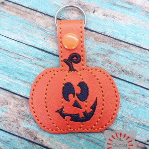 May include: Orange leather pumpkin keychain with a black stitched smiling face. The keychain has a silver metal ring.