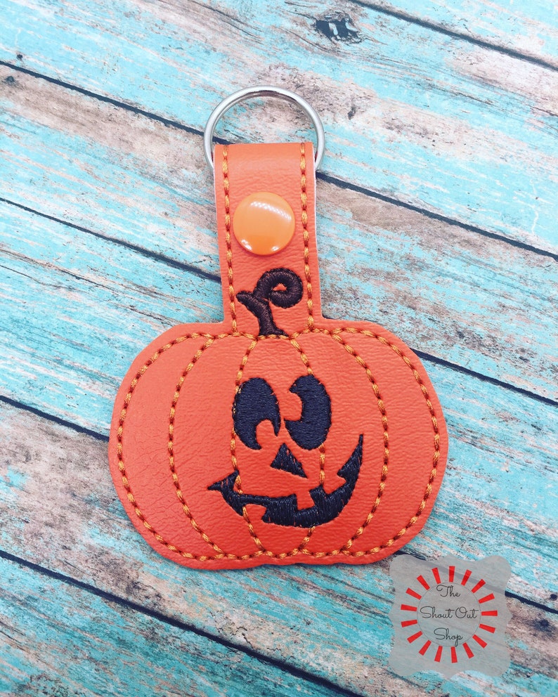 May include: Orange leather pumpkin keychain with a black stitched smiling face. The keychain has a silver metal ring.