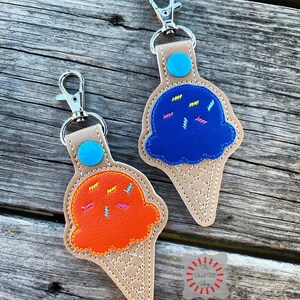 Puede incluir: Dos llaveros de cono de helado, uno azul y otro naranja, con chispas. Los llaveros est&aacute;n hechos de cuero y tienen un cono marr&oacute;n con un patr&oacute;n cosido blanco. Los llaveros tienen un c&iacute;rculo azul y turquesa en la parte superior y un cierre de metal plateado.
