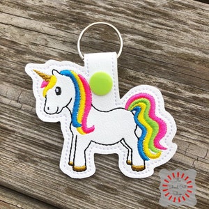 May include: White unicorn keychain with a rainbow mane and tail. The unicorn has a gold horn and green eyes. The keychain is attached to a silver key ring.