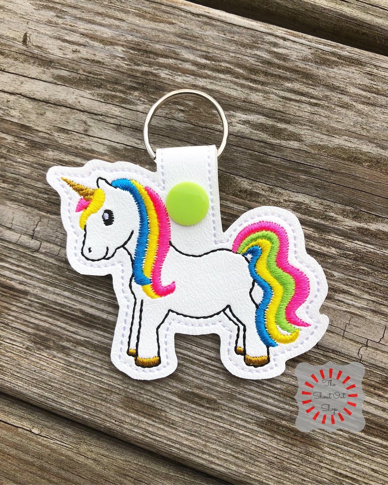 May include: White unicorn keychain with a rainbow mane and tail. The unicorn has a gold horn and green eyes. The keychain is attached to a silver key ring.