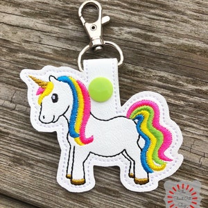 May include: A white unicorn keychain with a rainbow mane and tail. The unicorn has a gold horn and a green button on its side. The keychain is attached to a silver metal ring.