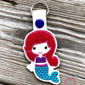 May include: A white fabric keychain with a blue button and a red-haired mermaid with a turquoise tail. The mermaid is smiling and has a purple top.