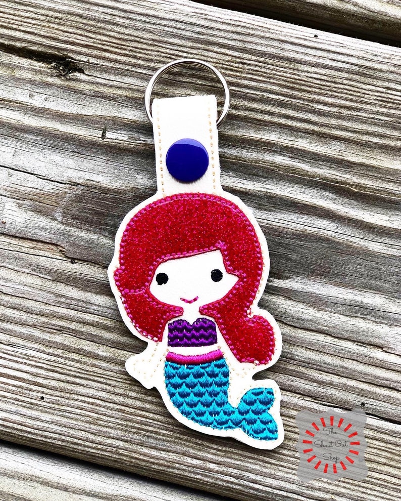 May include: A white fabric keychain with a blue button and a red-haired mermaid with a turquoise tail. The mermaid is smiling and has a purple top.