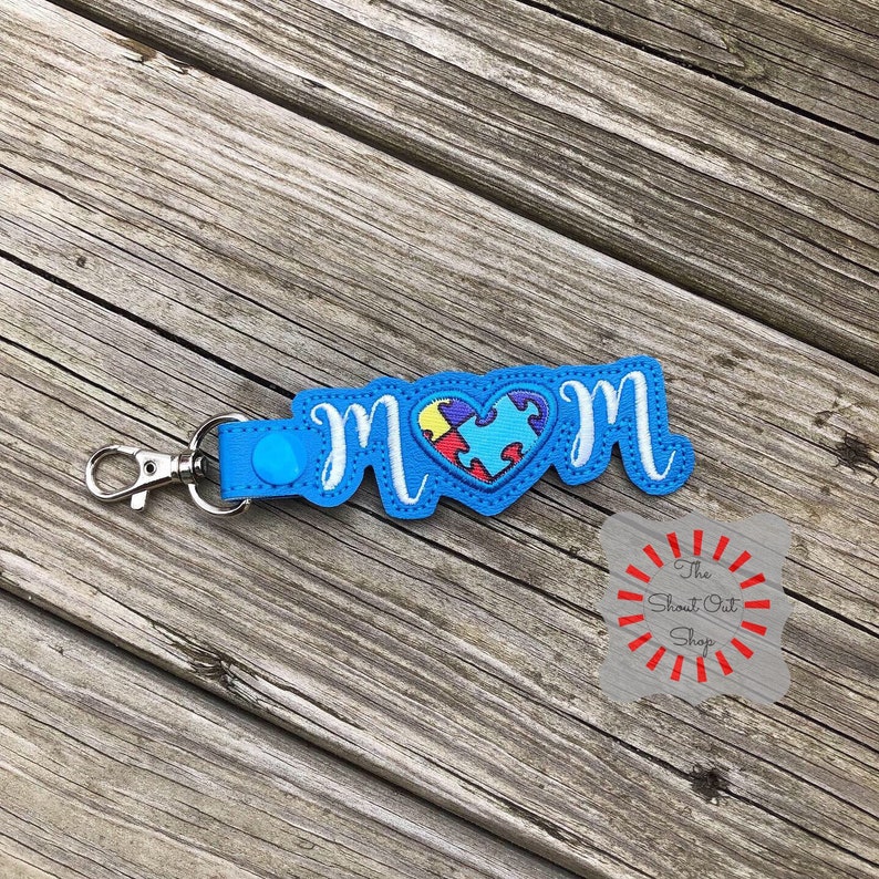 May include: Blue keychain with the word "MOM" in white cursive lettering and a blue and red puzzle piece heart in the center. The keychain has a silver metal clasp.
