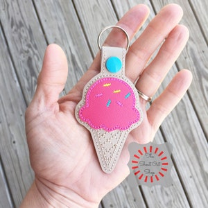 Puede incluir: Llavero de cono de helado rosa con chispas, bot&oacute;n azul y una correa de cuero beige. El llavero est&aacute; en una mano. El texto "The Shout Out Shop" est&aacute; en un c&iacute;rculo rojo con rayos blancos.