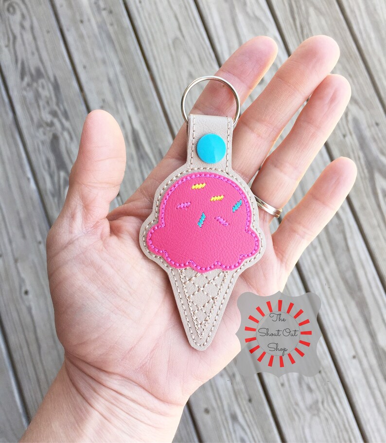 Puede incluir: Llavero de cono de helado rosa con chispas, bot&oacute;n azul y una correa de cuero beige. El llavero est&aacute; en una mano. El texto "The Shout Out Shop" est&aacute; en un c&iacute;rculo rojo con rayos blancos.