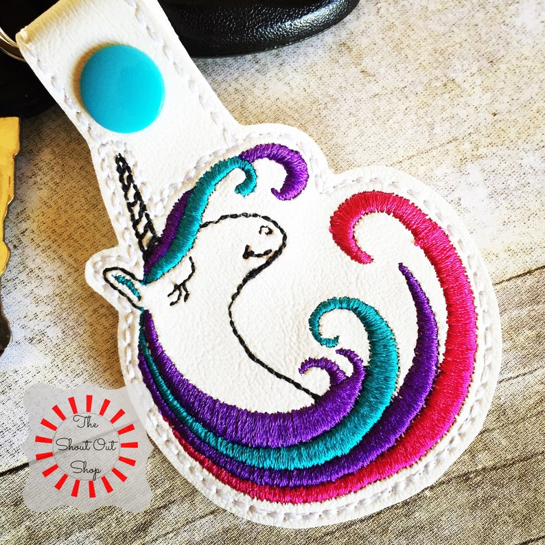 May include: White leather keychain with a colorful embroidered unicorn design. The unicorn has a purple, teal, and pink mane and tail. The keychain has a turquoise button.