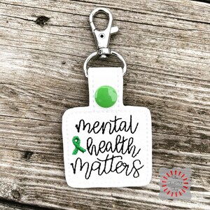 May include: White square keychain with a green ribbon and the text "mental health matters" in black cursive lettering. The keychain has a silver clip and a green button.