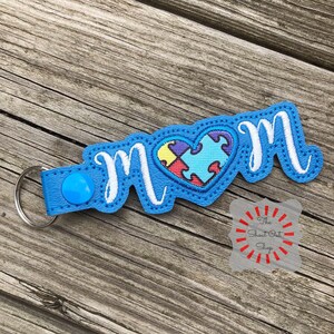 May include: Blue leather keychain with a white embroidered "MOM" and a heart-shaped puzzle piece design in the center. The heart is filled with colorful puzzle pieces.